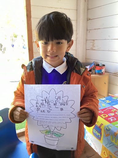 A young boy from Nursery is pictured holding up a sheet of paper with information he has found out about Sunflowers, whilst smiling for the camera.